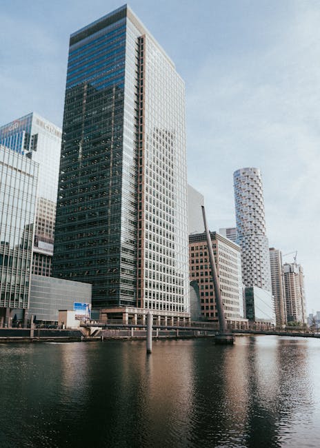 Photograph of modern high-rise office buildings in Canary Wharf, situated along the river Thames with clear blue sky overhead. The buildings feature glass facades reflecting sunlight, with one prominent black and white skyscraper at the centre, and surrounding structures including a cylindrical tower with a patterned exterior and other rectangular office blocks. In the foreground, the river Thames is visible with calm water and a few small boats moored along the dock. The scene captures the urban landscape of South Quay flats and commercial properties, relevant to house and furniture removals, with no interior elements visible. The image highlights the environment where professional removals by Man and a Van Canary Wharf may take place, focusing on the city’s distinctive architecture and waterfront setting.