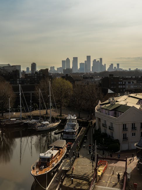 An urban scene showing a narrow waterway with several moored boats, including a wooden boat and a motorboat, along a paved waterfront area. To the right, there is a white residential building with large windows and a balcony, adjacent to a sidewalk where a few pedestrians are walking. In the background, a city skyline with high-rise buildings and skyscrapers under a partly cloudy sky is visible. The area appears to be part of a home relocation or furniture transport process, with the environment indicating an active moving or loading zone near Canary Wharf. The photograph captures a moment of loading or unloading activities associated with house removals, enhanced by the presence of environmental elements such as boats, packaging materials, and cityscape, consistent with professional moving services like those offered by Man and a Van Canary Wharf.