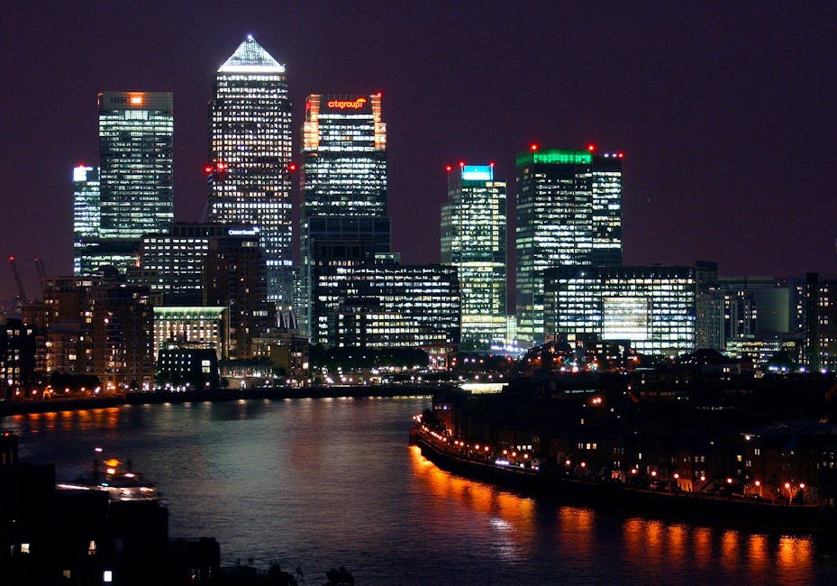 Photograph of modern high-rise office buildings in Canary Wharf, situated along the river Thames with clear blue sky overhead. The buildings feature glass facades reflecting sunlight, with one prominent black and white skyscraper at the centre, and surrounding structures including a cylindrical tower with a patterned exterior and other rectangular office blocks. In the foreground, the river Thames is visible with calm water and a few small boats moored along the dock. The scene captures the urban landscape of South Quay flats and commercial properties, relevant to house and furniture removals, with no interior elements visible. The image highlights the environment where professional removals by Man and a Van Canary Wharf may take place, focusing on the city’s distinctive architecture and waterfront setting.