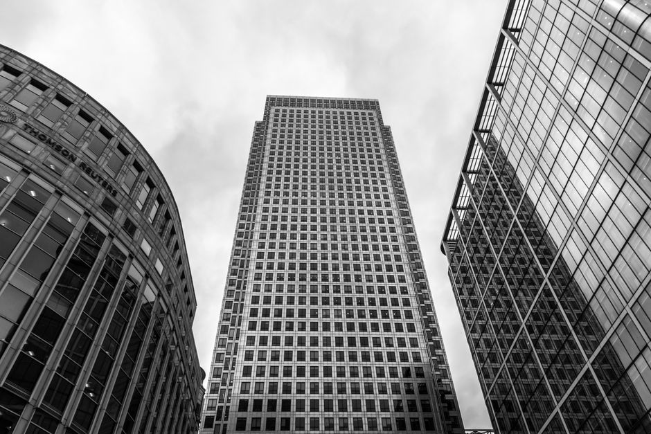 A clear daytime view of Canary Wharf featuring several modern high-rise office buildings with reflective glass facades and a waterway running along the base. In the foreground, a black metal railing l